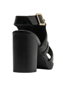 Elegante sandalia de cuero negro con acabado brillante, perforaciones y hebilla dorada.